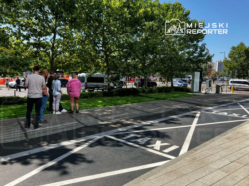 Grupa osób stoi na chodniku obok parkingu dla taksówek, w tle widać pojazdy i zieleń drzew.