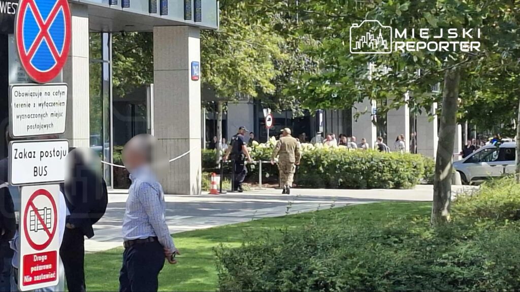 Grupa ludzi obserwująca interwencję służb mundurowych przed nowoczesnym biurowcem w słoneczny dzień.