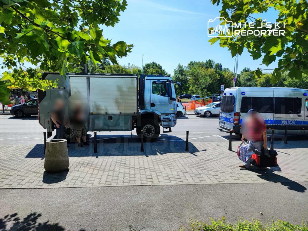 Mężczyzna z torbami siedzi na chodniku obok policyjnego auta oraz ciężarówki, w tle widać przechodniów.
