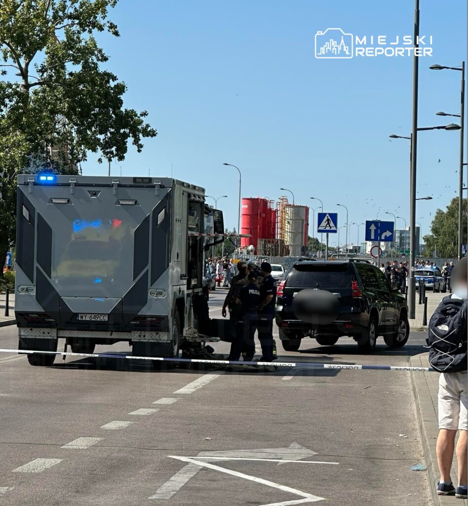 Policyjny wóz z włączonymi sygnałami świetlnymi stoi na ulicy, obok dwóch funkcjonariuszy badających miejsce zdarzenia.