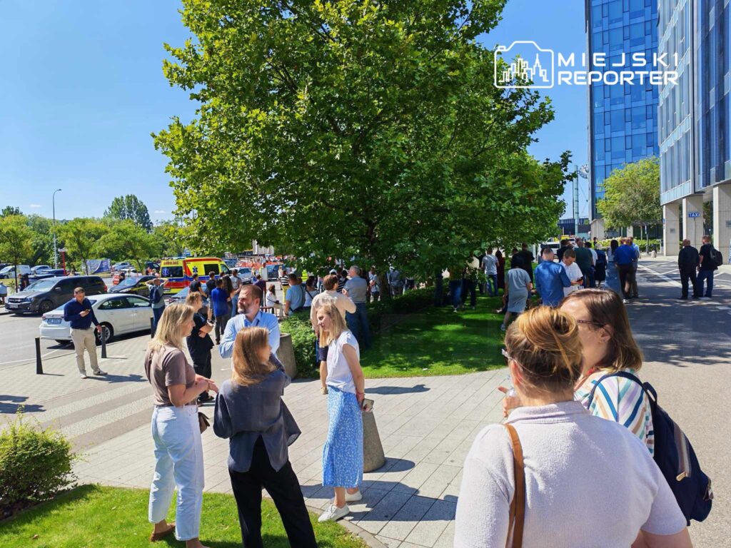Grupa ludzi w różnorodnych strojach rozmawia na chodniku w pobliżu nowoczesnego budynku biurowego w słoneczny dzień.