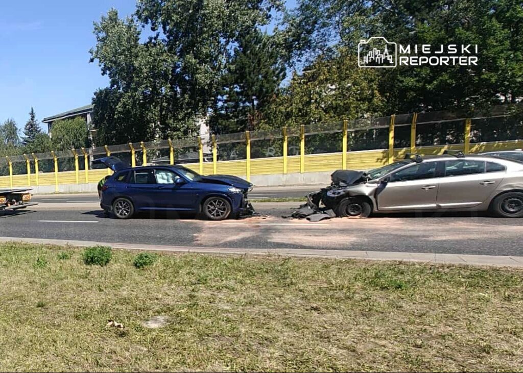 Dwa uszkodzone samochody osobowe stoją na drodze, obok żółta ogrodzenie i zieleń trawy.