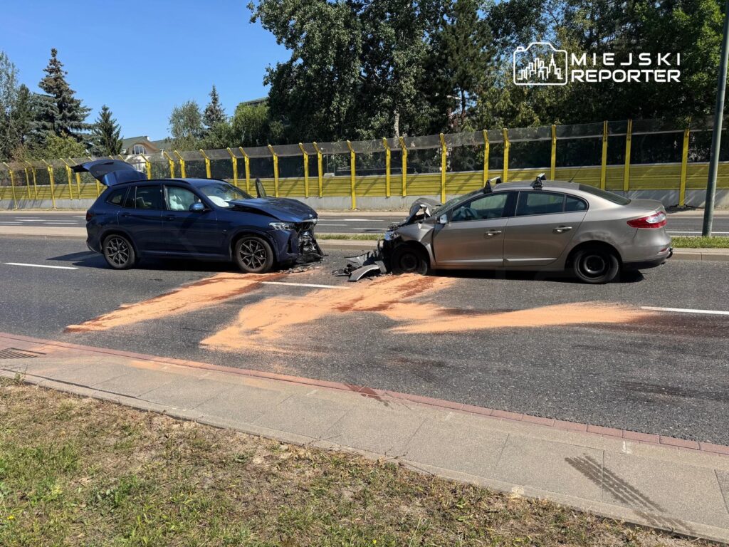 Dwa uszkodzone samochody osobowe stoją na jezdni, obok rozlanego piasku. W tle widoczna ogrodzenie i zieleń.