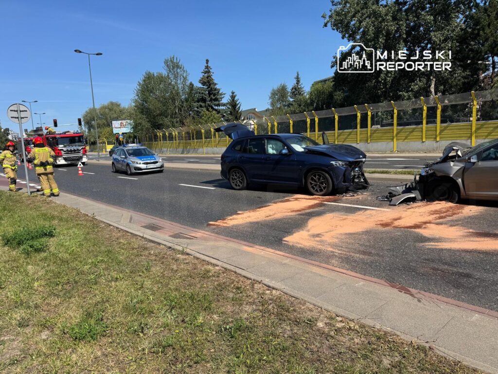 Dwa uszkodzone samochody na jezdni, obok stoją strażacy i policjanci, w tle widoczny ruch uliczny.