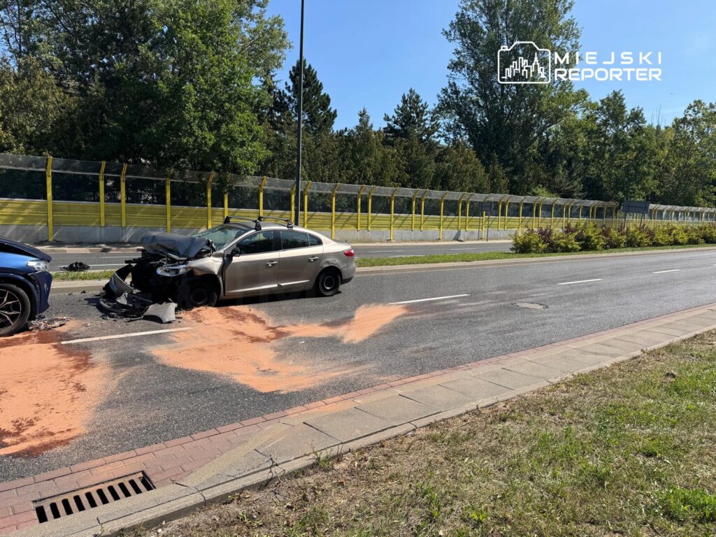 Uszkodzony samochód osobowy leży na jezdni obok drugiego pojazdu, a na drodze widoczny jest ślad czerwonego proszku.