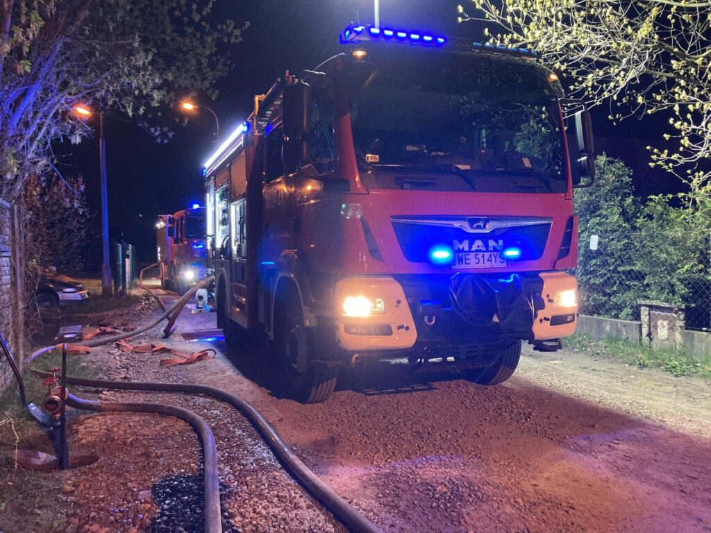 Czerwony wóz strażacki z niebieskimi światłami stoi na wąskiej drodze nocą, obok leżą węże pożarnicze.