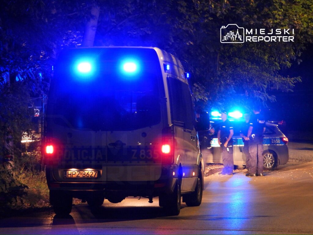Nocna akcja policji z niebieskimi światłami, funkcjonariusze rozmawiają przy radiowozie na oświetlonej ulicy.