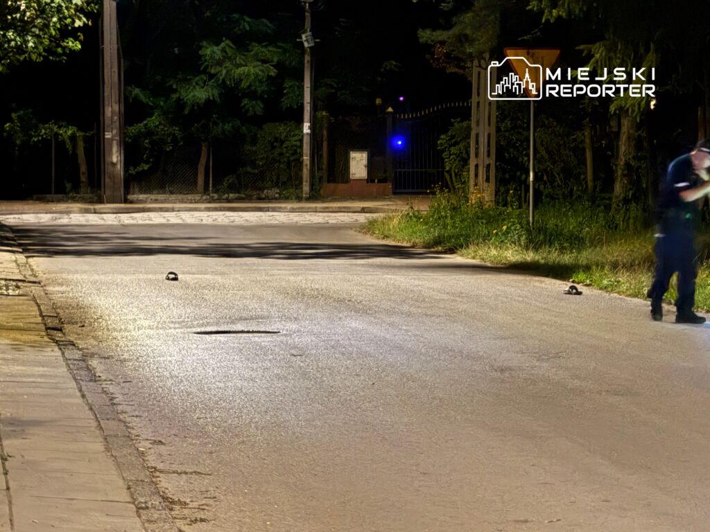 Policjant bada miejsce zdarzenia na oświetlonej ulicy, obok leżą dwa przedmioty na jezdni.