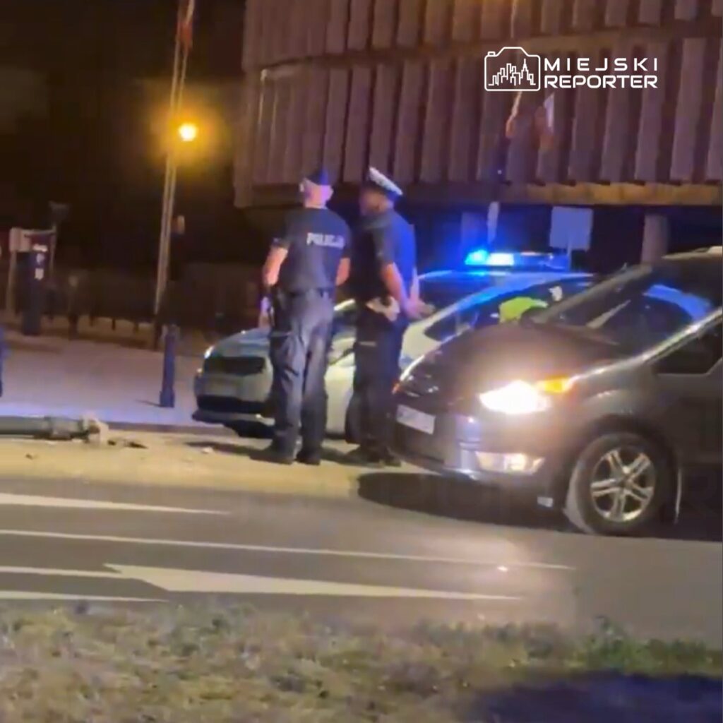 Dwaj policjanci w mundurach rozmawiają obok radiowozu na oświetlonej ulicy nocą.