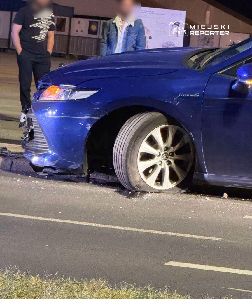 Niebieski samochód osobowy z uszkodzoną oponą stoi na drodze, obok dwóch mężczyzn rozmawiających ze sobą.