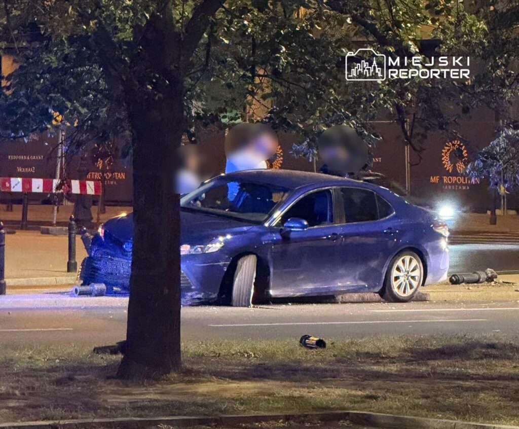 Niebieski samochód osobowy z uszkodzoną oponą zaparkowany na ulicy, obok leżące przedmioty. W tle widoczne drzewa i oświetlenie nocne.