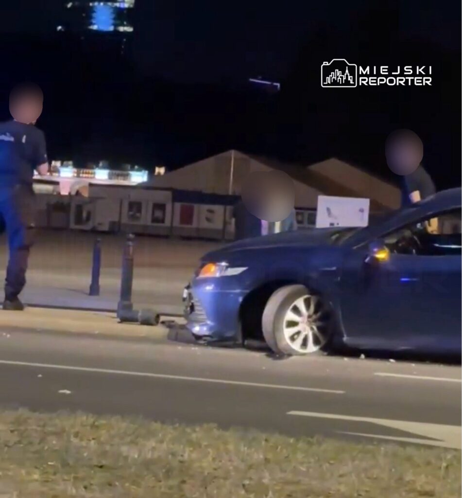 Dwaj policjanci rozmawiają obok samochodu osobowego z uszkodzoną oponą na nocnej ulicy.