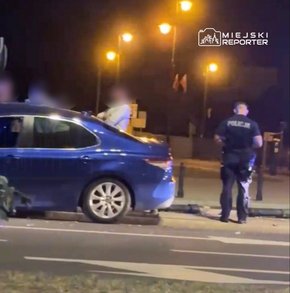 Mężczyzna w białej koszuli stoi na dachu niebieskiego samochodu, podczas gdy policjant rozmawia z innym mężczyzną obok pojazdu. Nocne oświetlenie uliczne w tle.
