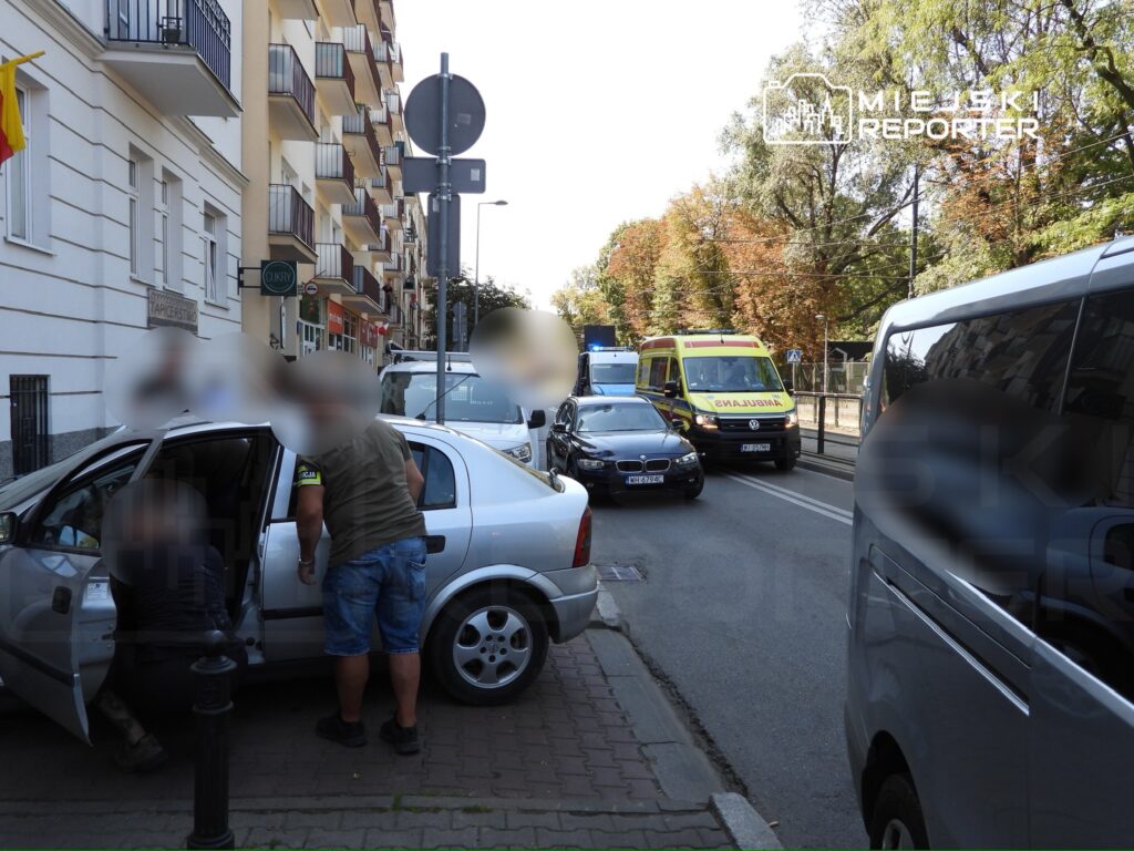 Mężczyzna w krótkich spodniach rozmawia z osobą w samochodzie na ruchliwej ulicy, obok przystanek autobusowy.