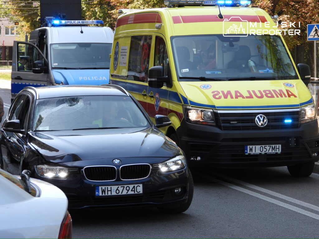Czarny samochód osobowy zatrzymany obok karetki pogotowia i radiowozu policyjnego na miejskiej ulicy.