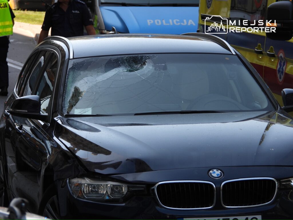 Uszkodzony czarny samochód osobowy z pękniętą szybą przednią stoi na drodze, obok policjanci i pojazdy służb ratunkowych.