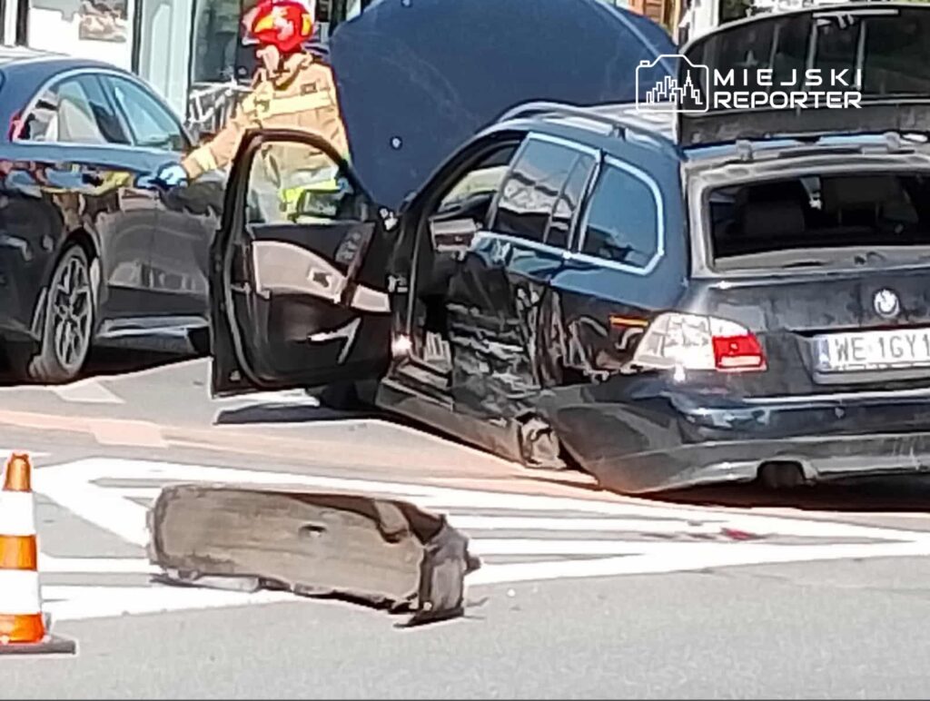Strażak w hełmie bada uszkodzony samochód osobowy na skrzyżowaniu, obok leży oderwana opona.