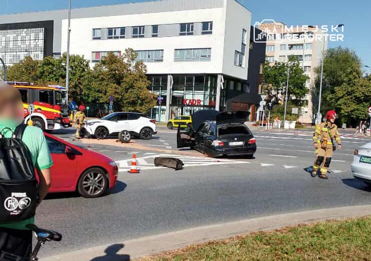 Na skrzyżowaniu stoi czarny samochód z otwartą klapą, obok niego pracują strażacy, a w tle widać nowoczesny biurowiec.