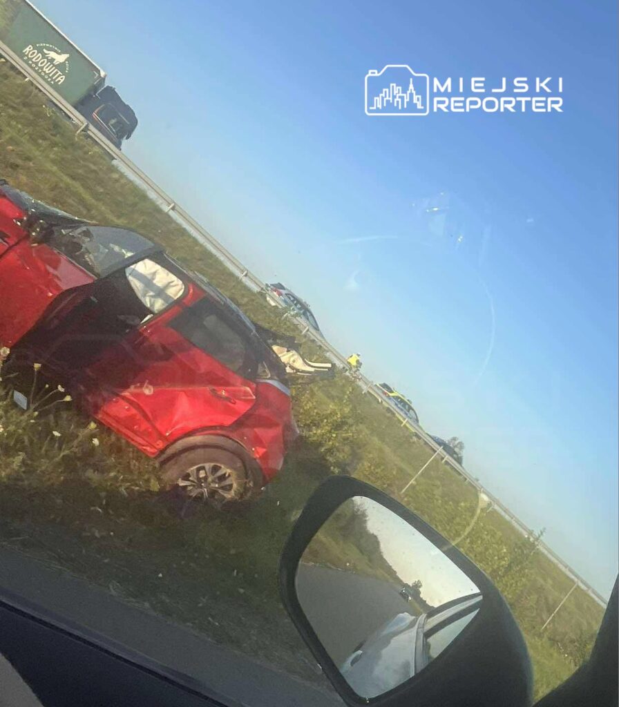 Zniszczony czerwony samochód osobowy leży w przydrożnym rowie, obok ruchliwej drogi, w tle widać policyjne pojazdy.