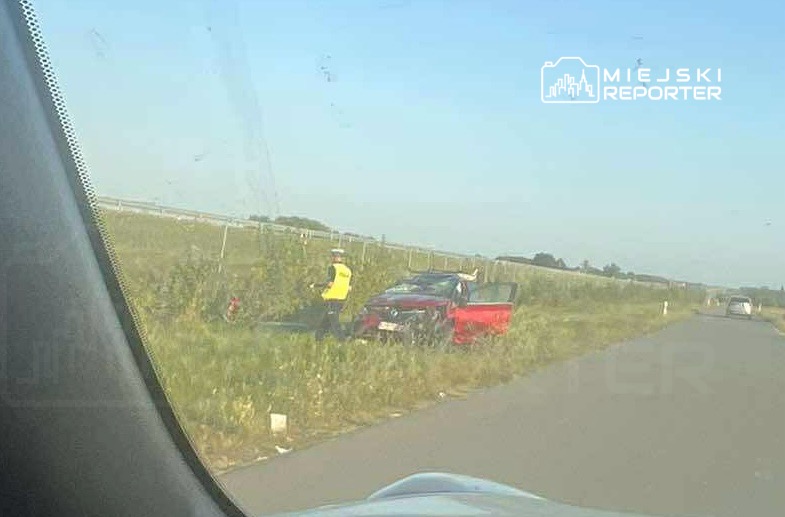 Mężczyzna w żółtej kamizelce sprawdza uszkodzony czerwony samochód na poboczu drogi w słoneczny dzień.