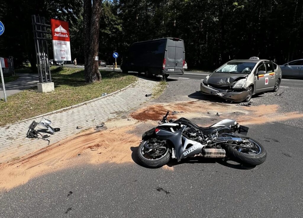 Motocykl leżący na drodze obok rozbitego samochodu osobowego, w tle znaki drogowe i zieleń.