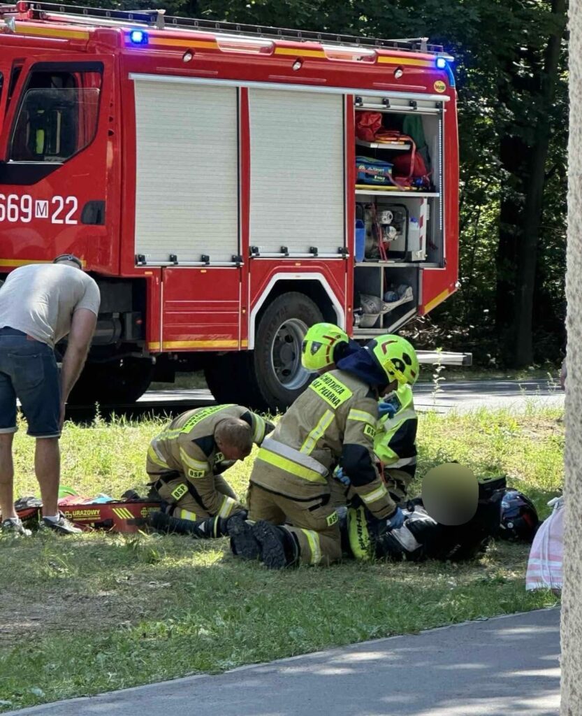 Grupa strażaków w mundurach udziela pomocy poszkodowanemu na trawie obok wozu strażackiego.