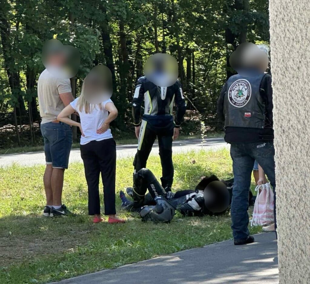 Grupa osób, w tym motocyklista w kombinezonie, obserwuje leżącego na ziemi mężczyznę przy drodze w otoczeniu drzew.