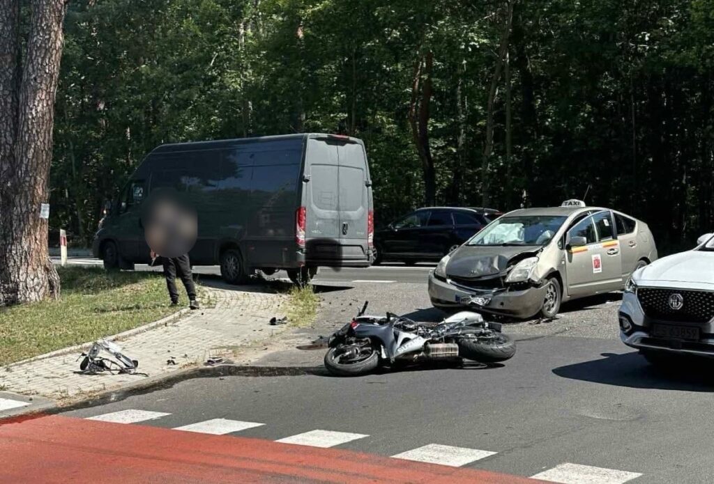 Motocykl leży na jezdni obok uszkodzonego taxi, a kierowca stoi na chodniku obok czarnego vana w lesistym otoczeniu.