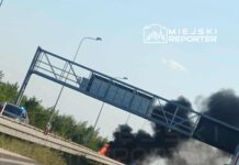 Pożar pojazdu na poboczu drogi, z dymem unoszącym się w powietrzu, obok policyjny radiowóz.