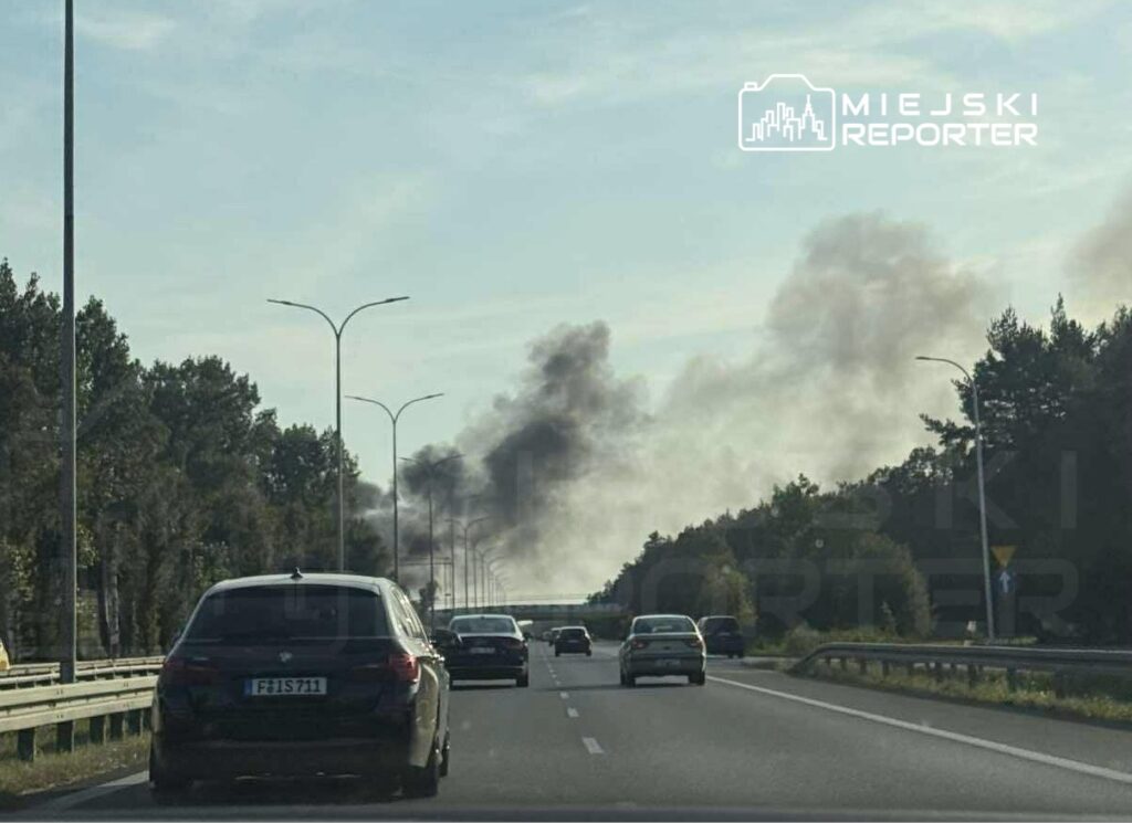 Na drodze widoczne są samochody, a w tle unosi się czarny dym z kierunku lasu.
