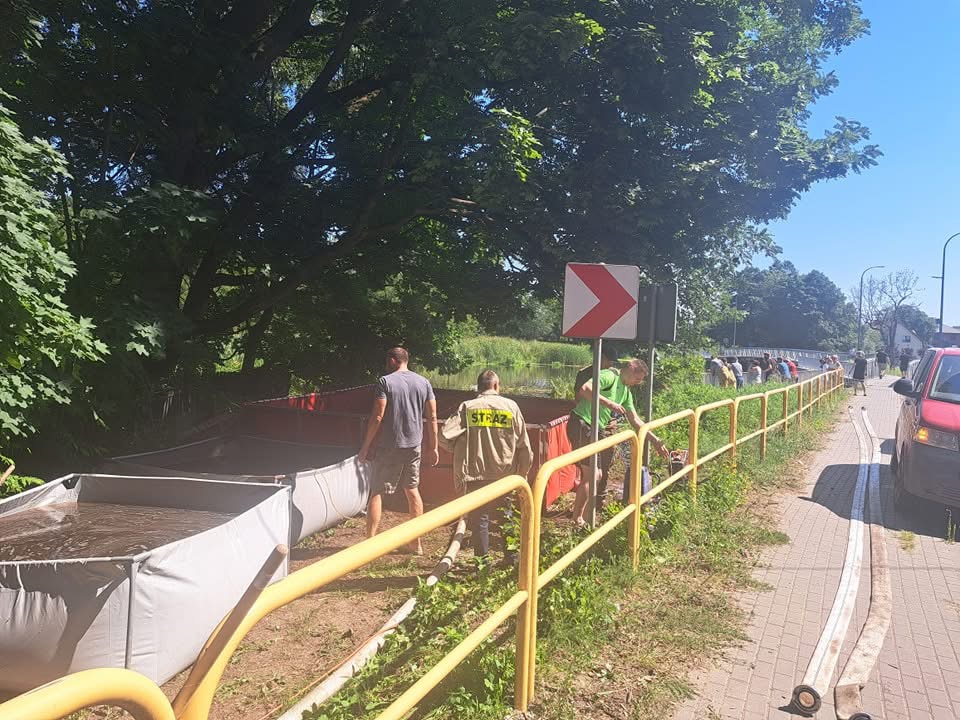 Grupa osób napełnia worki z piaskiem wzdłuż rzeki, obok żółtej bariery ochronnej, w słoneczny dzień.