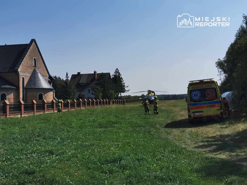 Grupa strażaków i ratowników medycznych pracuje na łące obok kościoła, w tle widoczna karetka pogotowia.