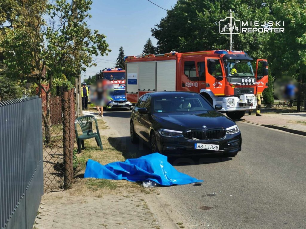 Na drodze leży niebieski materiał, obok czarny samochód osobowy oraz pojazdy straży pożarnej i policji.