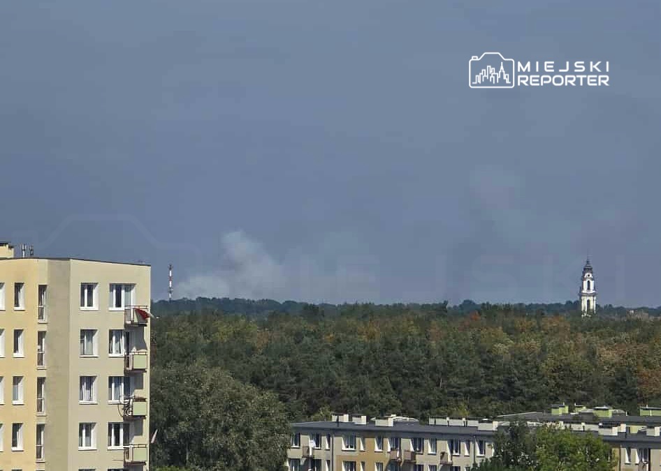Widok na nowoczesny budynek mieszkalny z balkonami, w tle dym unoszący się nad lasem oraz wieżą kościoła.