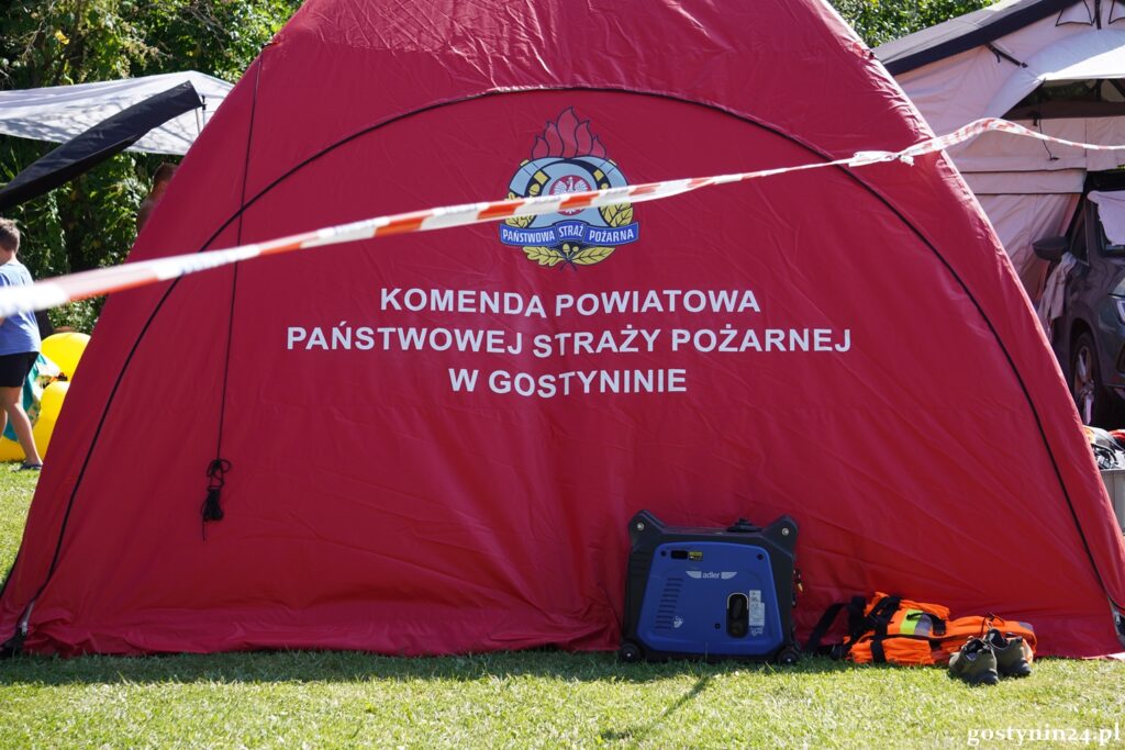 Czerwony namiot z logo straży pożarnej, obok generator prądu i pomarańczowe akcesoria na trawie.