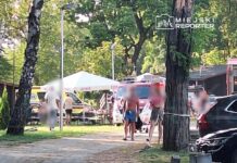 Grupa ludzi w letnich ubraniach przebywa w parku, obok namiotu z napisem "TYSIĄC". W tle widoczne karetki pogotowia i samochody.