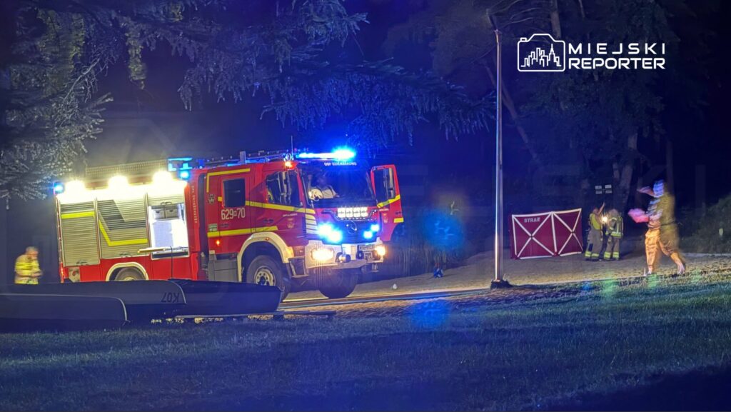 Czerwony wóz strażacki z włączonymi sygnałami świetlnymi stoi na drodze, obok dwóch strażaków w mundurach.