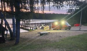 Grupa ratowników pracuje przy brzegu jeziora, obok karetki z włączonymi światłami. W tle widać pomost.