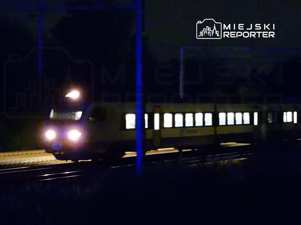 Pociąg osobowy z włączonymi światłami przejeżdża po torach w ciemnej scenerii nocnej.