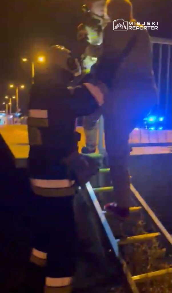 Strażak w mundurze pomaga mężczyźnie zejść z drabiny w nocy, otoczeni przez oświetlenie uliczne.