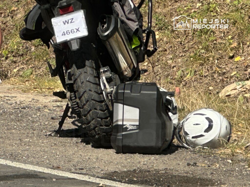 Motocykl z dużym kołem stoi obok torby i kasku na poboczu drogi w otoczeniu trawy i kamieni.
