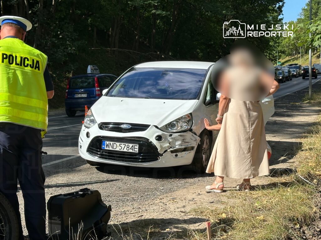 Policjant w kamizelce odblaskowej rozmawia z kobietą obok uszkodzonego białego samochodu na drodze.
