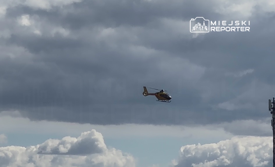 Helikopter w żółto-szarej kolorystyce przelatuje nad chmurami w jasnoniebieskim niebie.