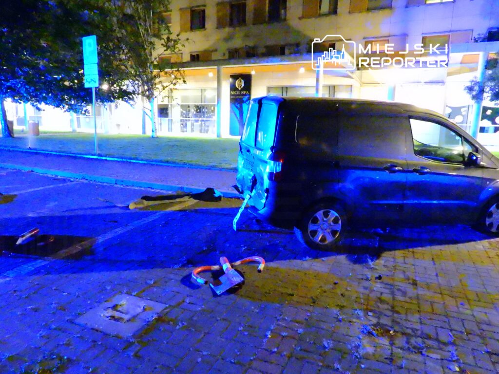 Czarny samochód dostawczy uszkodzony na ulicy, obok leżące elementy drogi i oświetlenie w niebieskim odcieniu.