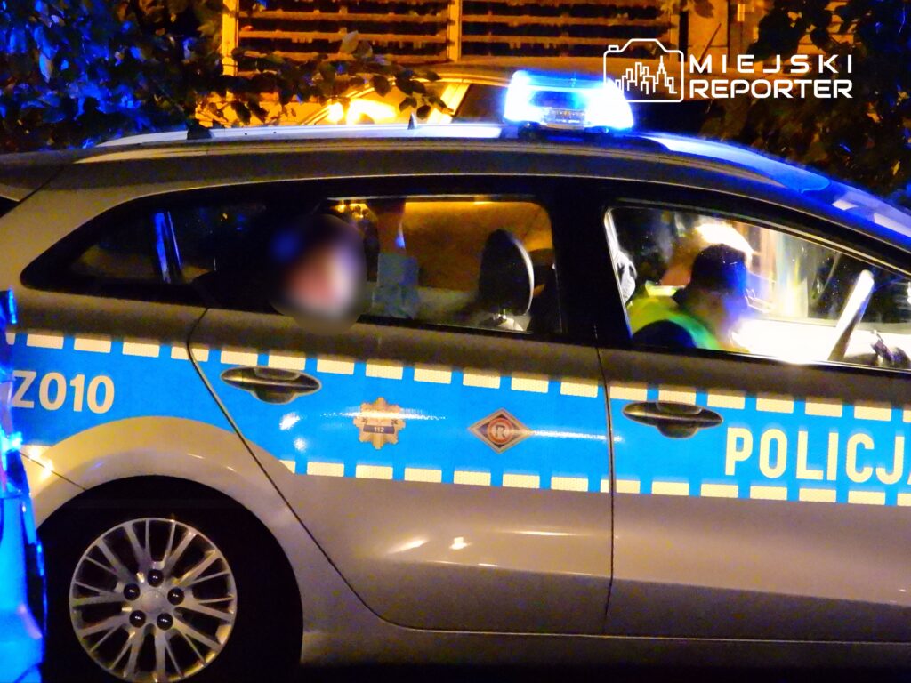 Policjant w radiowozie z niebieskimi światłami, rozmawiający przez radio w nocy.