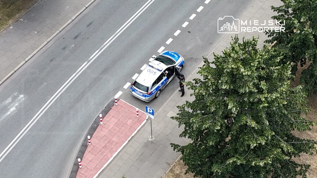 Policjant wysiada z radiowozu zaparkowanego na ulicy obok chodnika, w otoczeniu drzew.