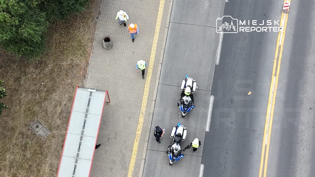 Grupa pracowników w kamizelkach odblaskowych oraz policjant na drodze obok zaparkowanych motocykli służbowych.