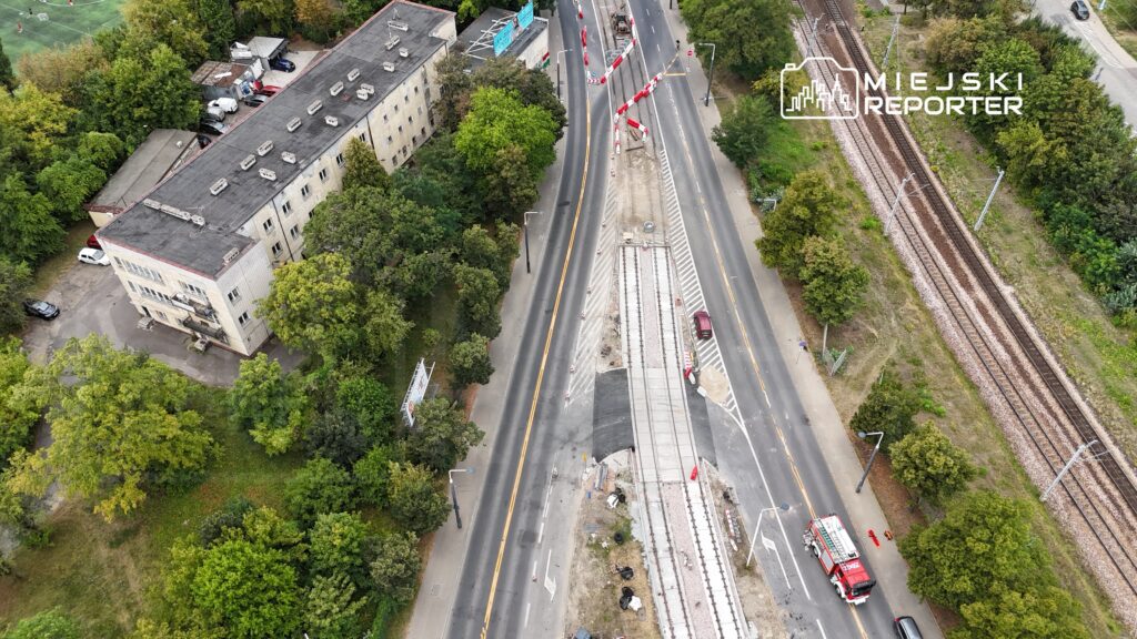 Widok z góry na ulicę z torami tramwajowymi w trakcie budowy, otoczoną drzewami i budynkami mieszkalnymi.