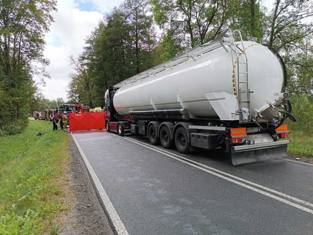 Ciężarówka z cysterną stoi na drodze, a strażacy zabezpieczają teren, ustawiając czerwoną barierę.