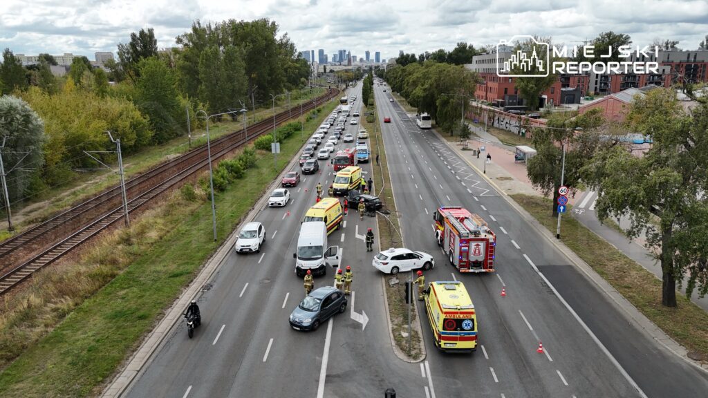 Na drodze widać wypadek samochodowy, obok stoją pojazdy służb ratunkowych oraz zablokowane samochody.